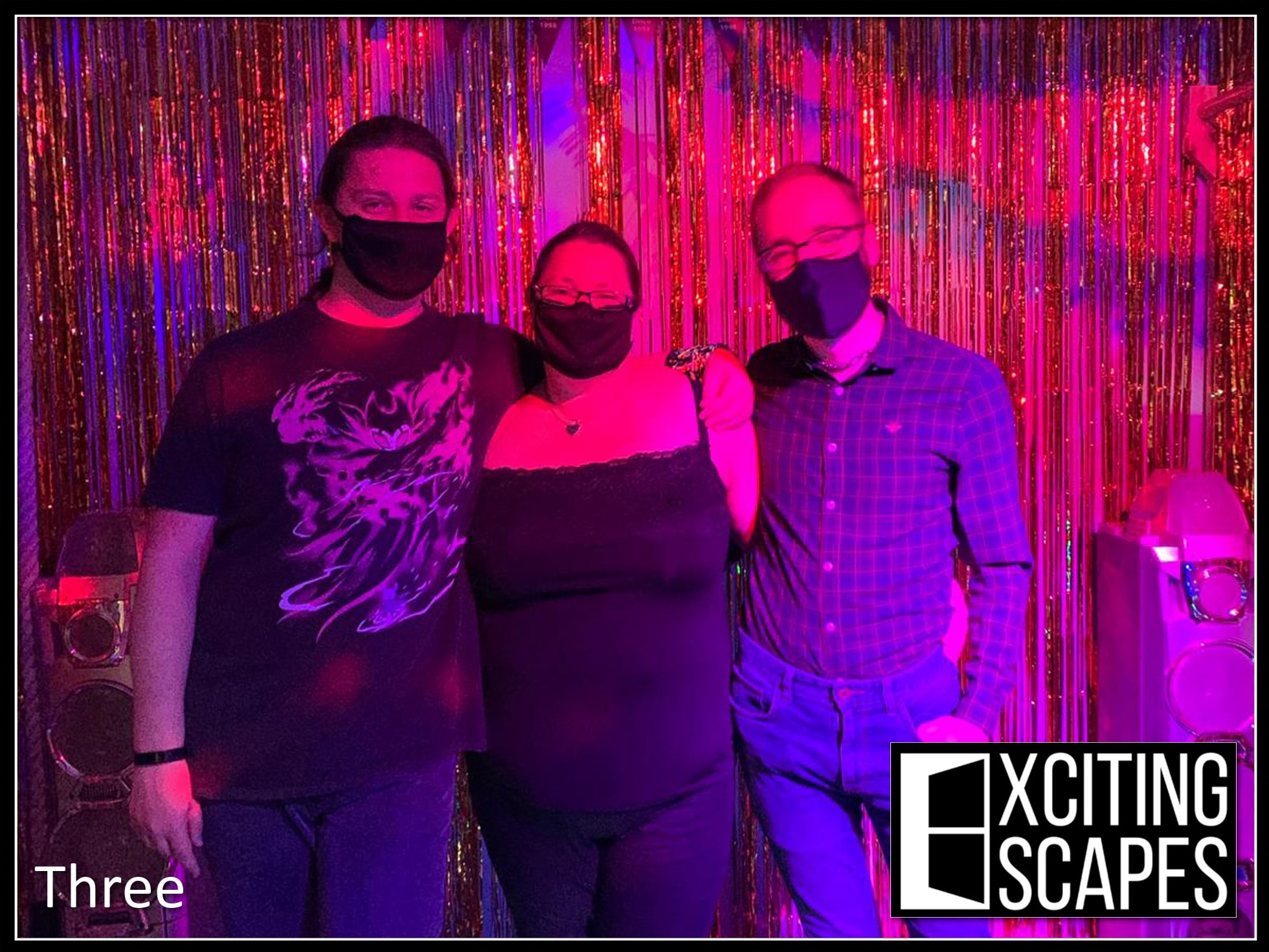 A three-person team pose after Danger on the dance floor (90's) at Exciting Escapes, before metallic party streamers in pink and purple light.