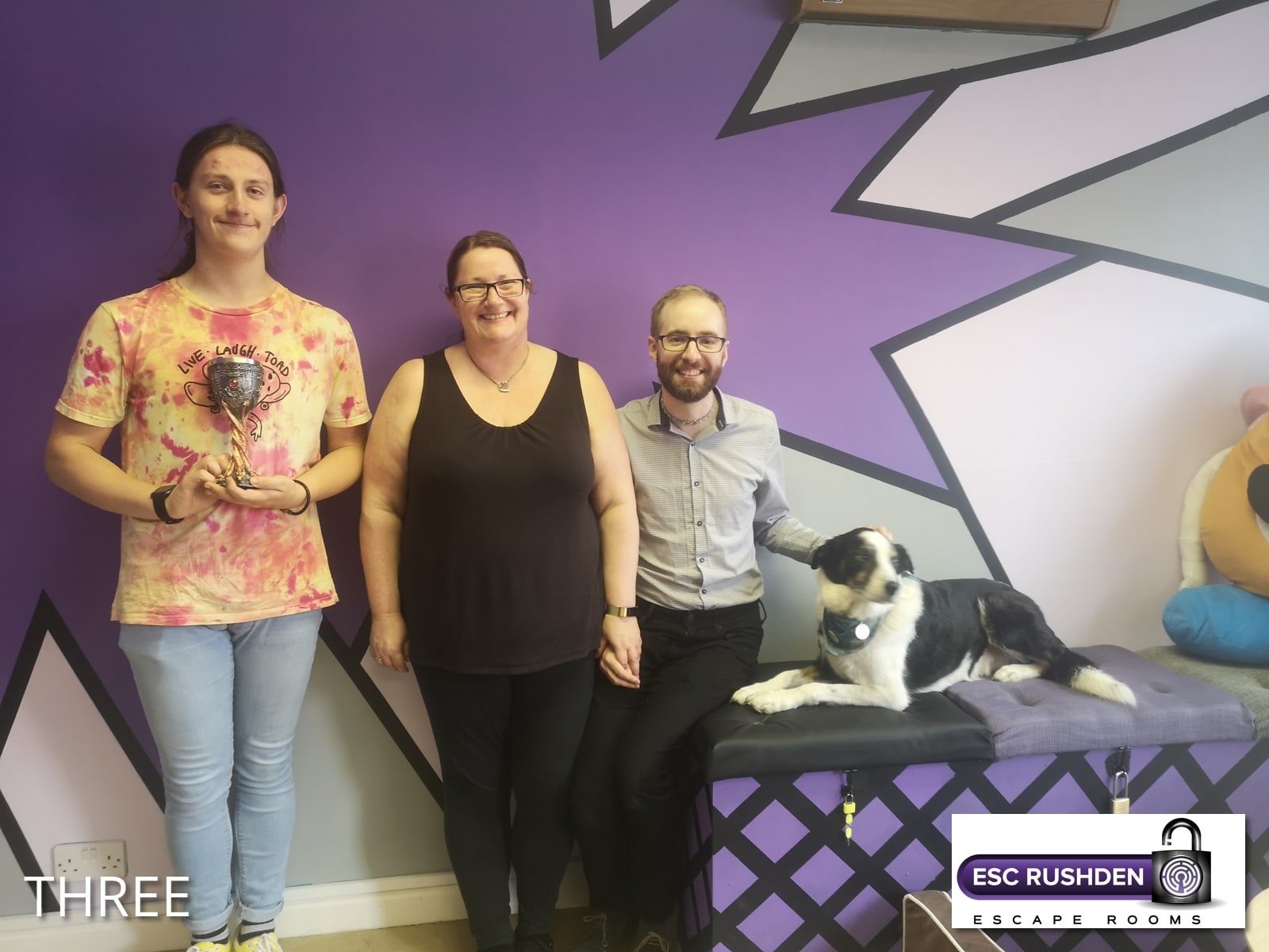 A three-person team pose after Guardian of the gallery at Escape Rushden, in the purple Escape Rushden room beside a dog.