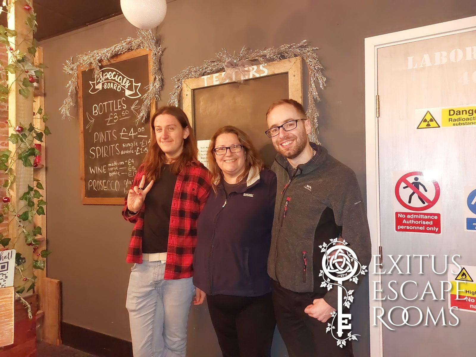 A three-person team pose after Coco Loco at Exitus, beside pub chalkboards and a laboratory warning door.